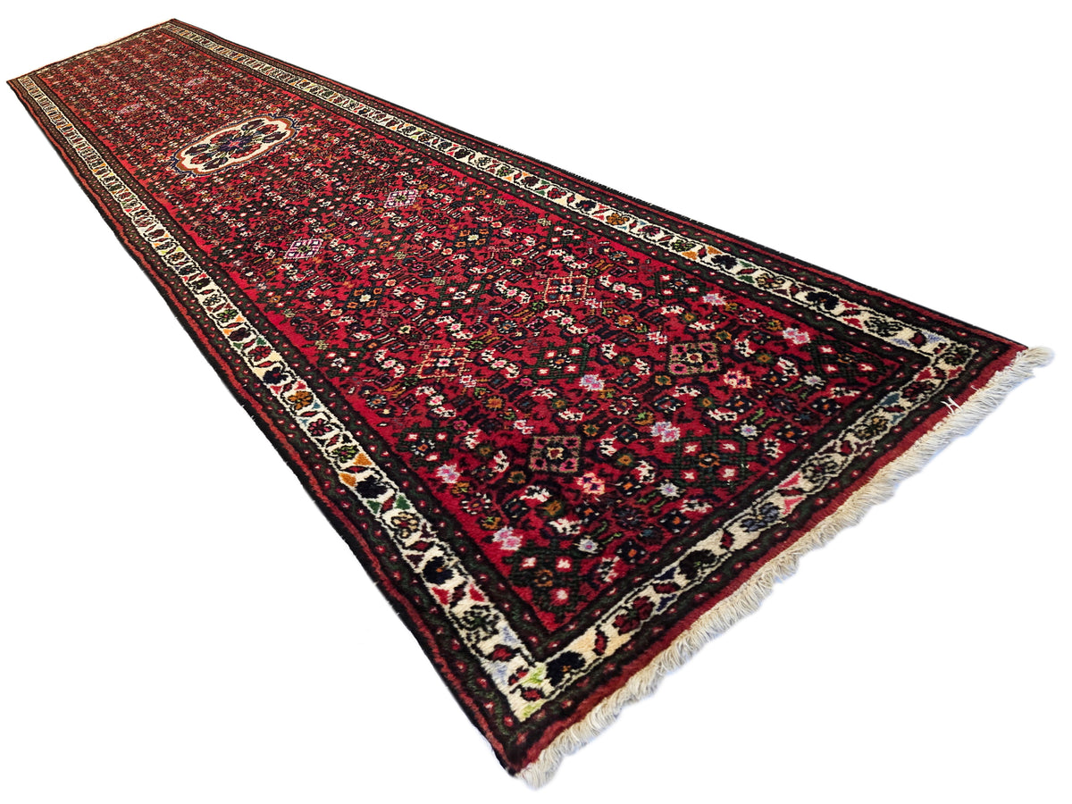 Red runner rug with dark floral medallion stripe, cream-and-black border, short fringe