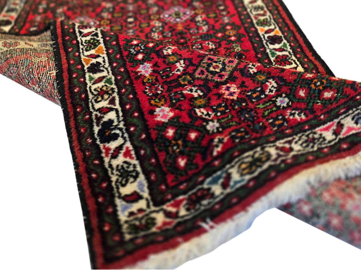 Red runner rug corner detail with dark floral motifs, cream border band, short white fringe