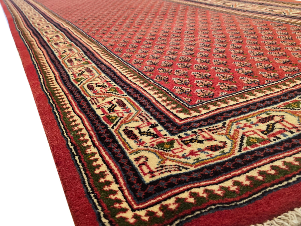 Red rug corner with cream floral motifs, dark border, and braided trim along edge