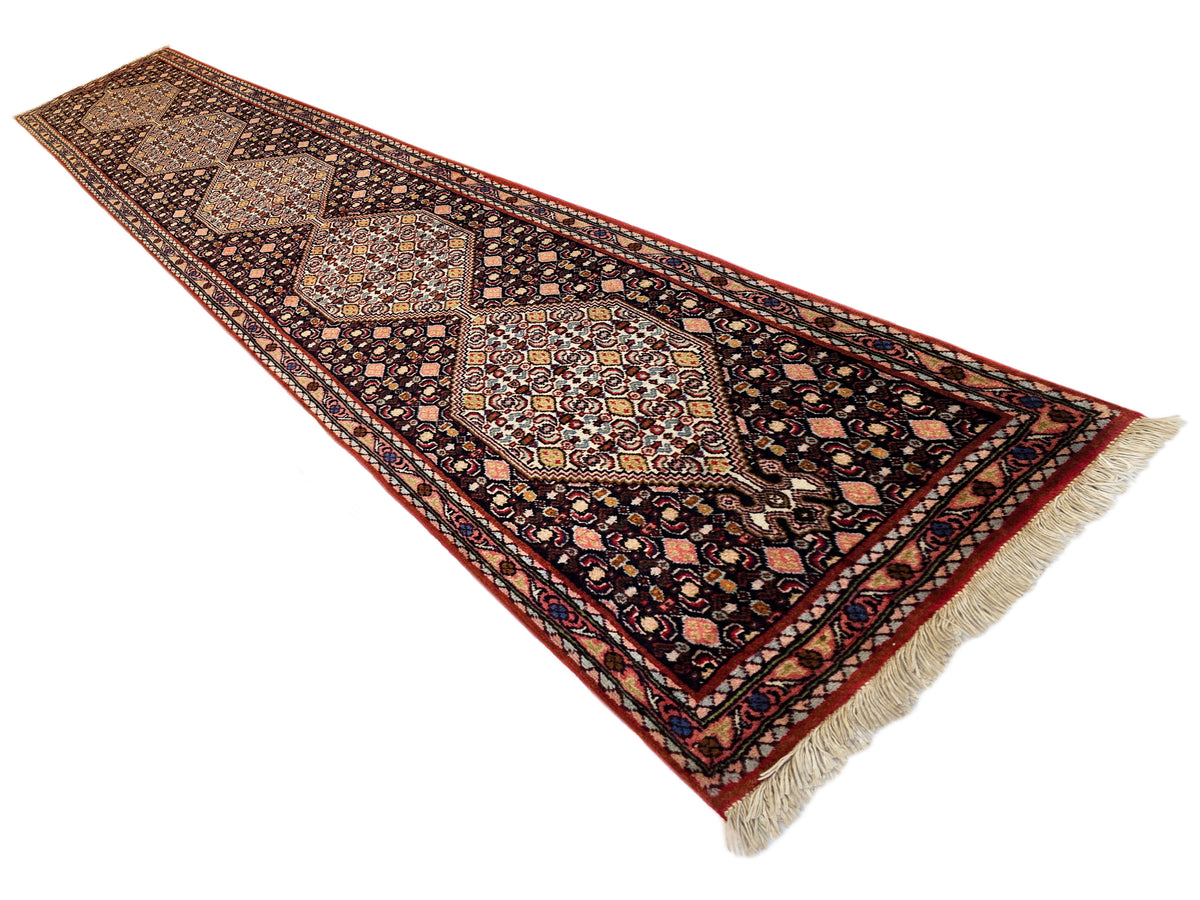 Diagonal view of narrow runner rug with dark field, red border, repeating geometric medallions, fringe.