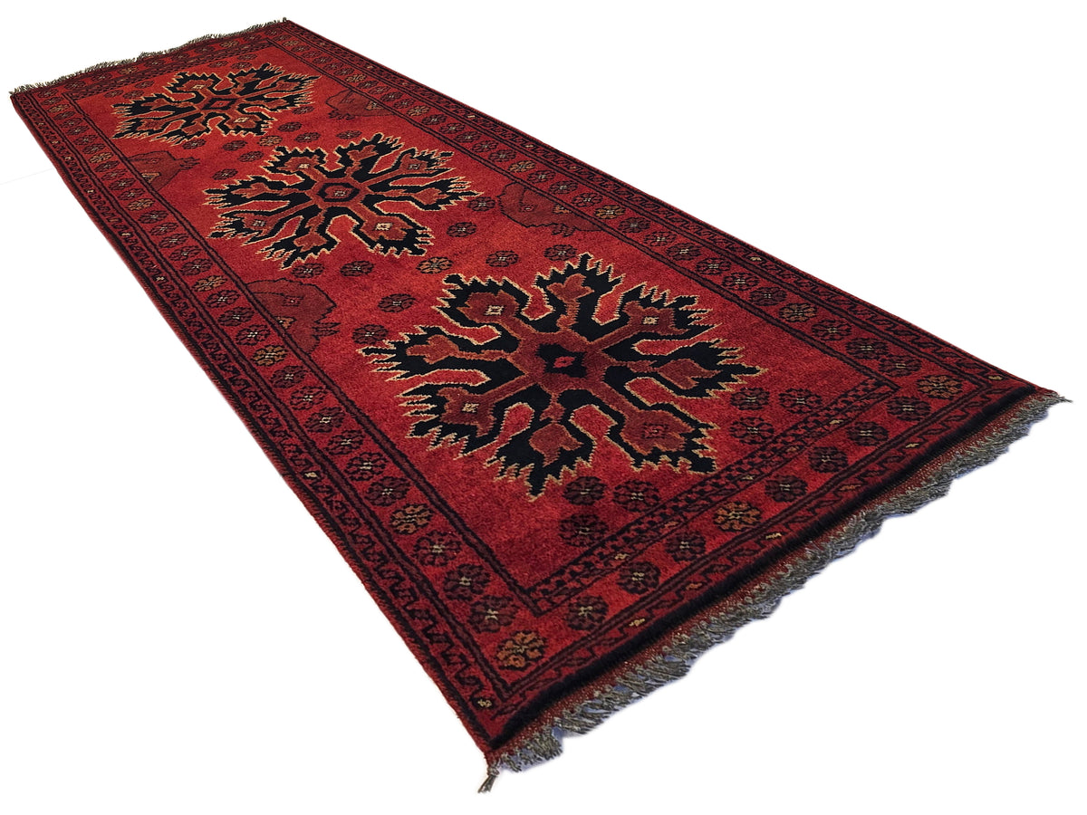 Red geometric Afghan rug with dark border, stylized motifs, fringed corners, 145x55 cm.