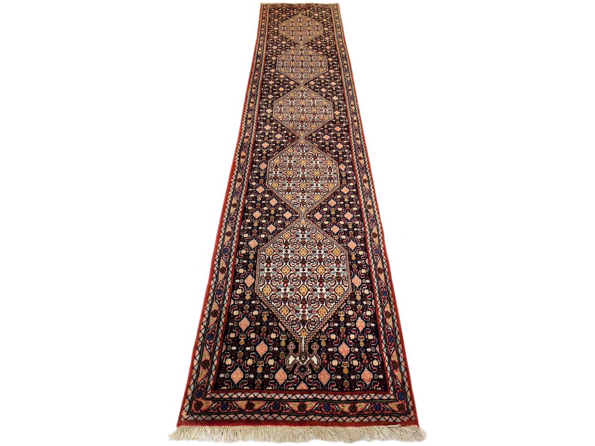 Narrow patterned runner rug with dark base, red border, diamond motifs, fringe ends.