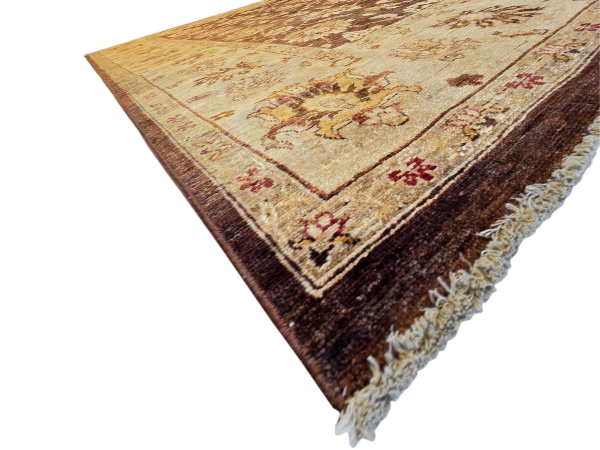 Corner view of ornate rug with tan top, dark brown field, and cream fringe